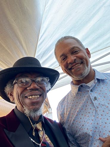 Two smiling older men posing for a selfie under a canopy.