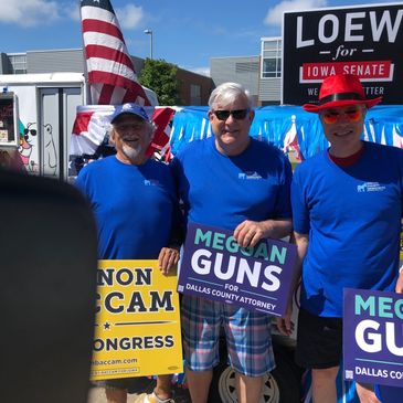 Three men in blue shirts holding political campaign signs at an outdoor event.