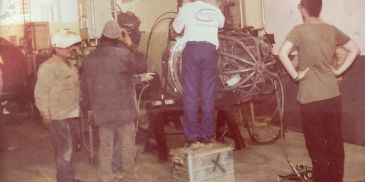 Workers operating machinery while one stands on a box.
