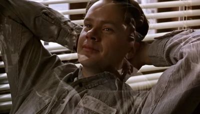 A man in a prison uniform leaning back with hands behind his head, looking contemplative.