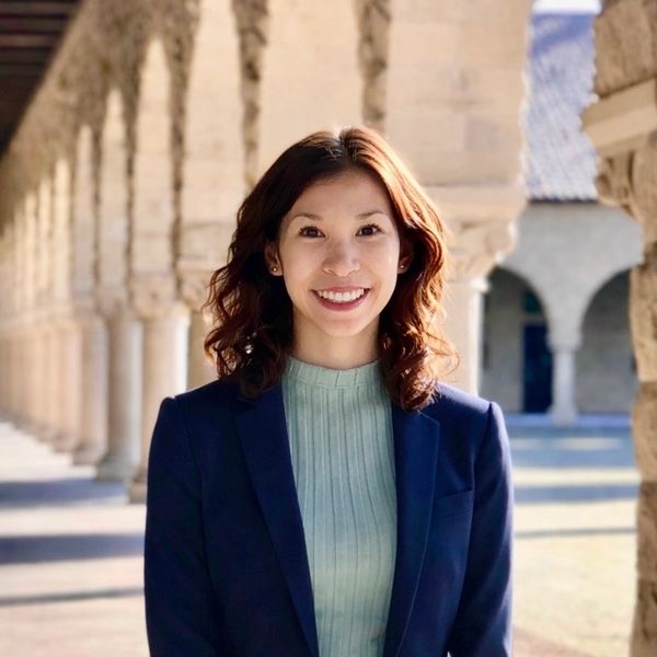 A woman smiling in a sunlit stone archway corridor.