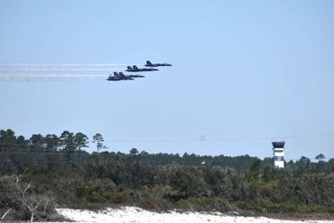 A formation of six jet planes flying in the sky above trees and a control tower.