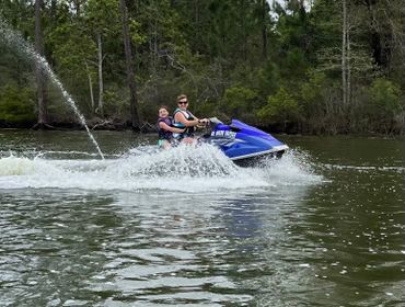 Two people riding a blue jet ski on a lake surrounded by trees.