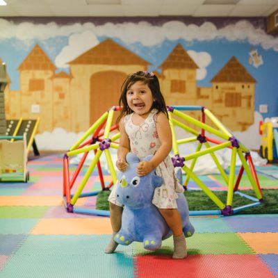 A young girl happily rides a blue plush rhino toy indoors.