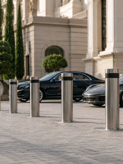 Metal bollards line a driveway with luxury cars and classical architecture in the background.