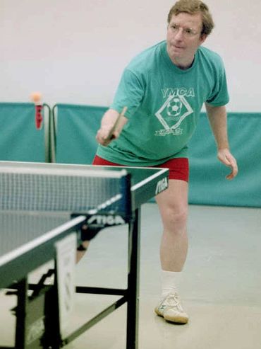 Man in green YMCA shirt plays table tennis indoors.