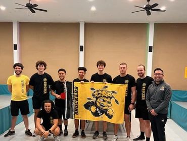 A group of young men posing with a Wichita State Shockers flag in a gym.