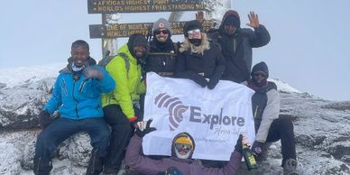 Group of climbers celebrating at Uhuru Peak, Mount Kilimanjaro, with snowy rocks and a sign.