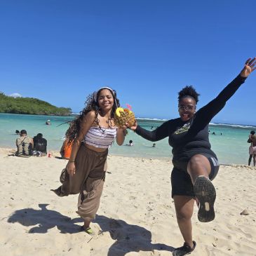 Two friends joyfully posing on a sunny beach with a pineapple drink.