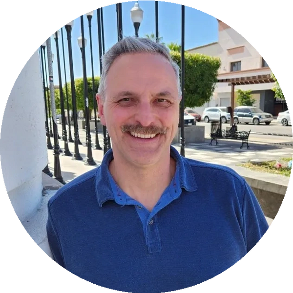 Smiling man with a mustache in a blue shirt outdoors on a sunny day.
