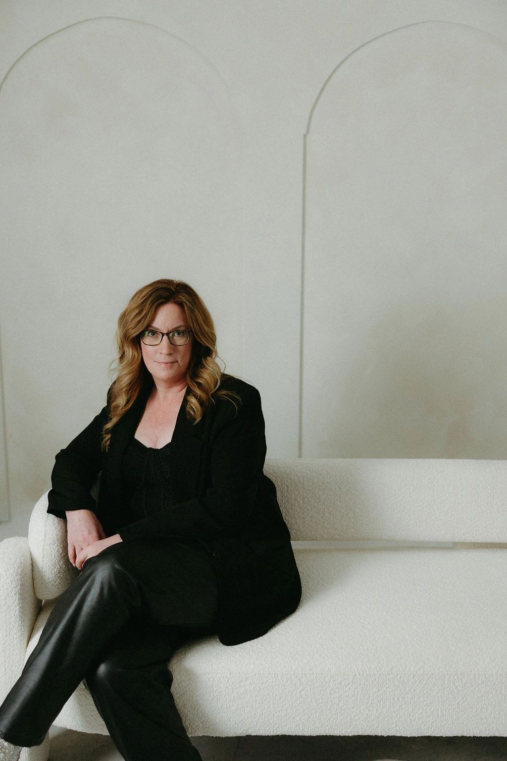 Confident woman in black outfit sitting on a white couch.