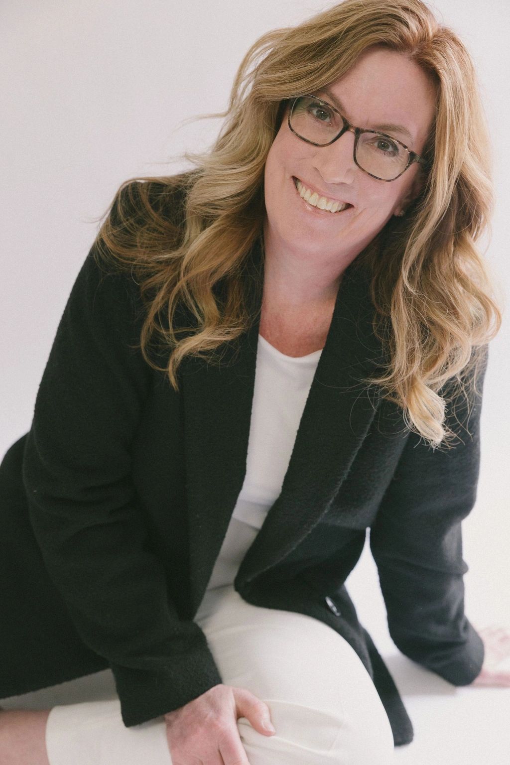 Smiling woman with glasses wearing a black jacket and white pants.
