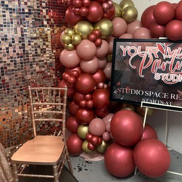 Rose gold-themed party setup with balloons, a chair, and a decorative sign.