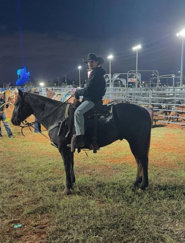 Man in cowboy hat rides a black horse at night in a rodeo arena.