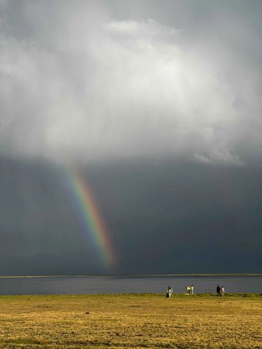 A rainbow appears over a lake with people standing on the shore.