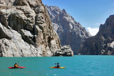 Two kayakers paddle on turquoise water surrounded by towering rocky mountains.