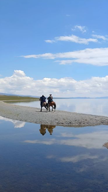 Two riders on horseback by a calm lakeside under a clear sky.