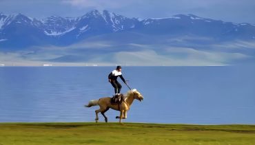 Man riding a horse by a lake with snow-capped mountains in the background.