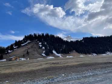 Mountain landscape with scattered snow and houses under a cloudy blue sky.
