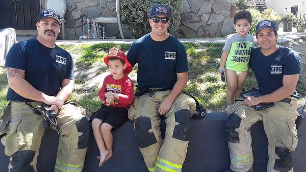 Two firefighters sit with two young boys on a grassy area, smiling and enjoying a sunny day.