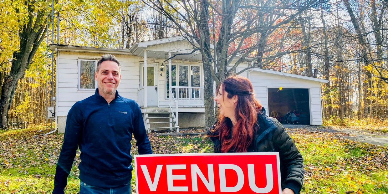 Happy couple holding a "VENDU" sign in front of their sold house on a sunny autumn day.