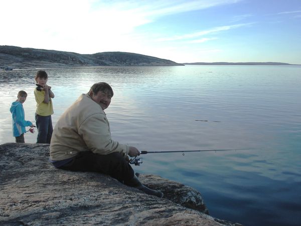 A man and two children fishing by a calm lake on rocky shore.