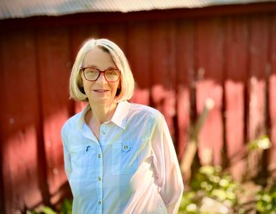 A woman with glasses stands outdoors in front of a red wooden wall, wearing a white shirt.