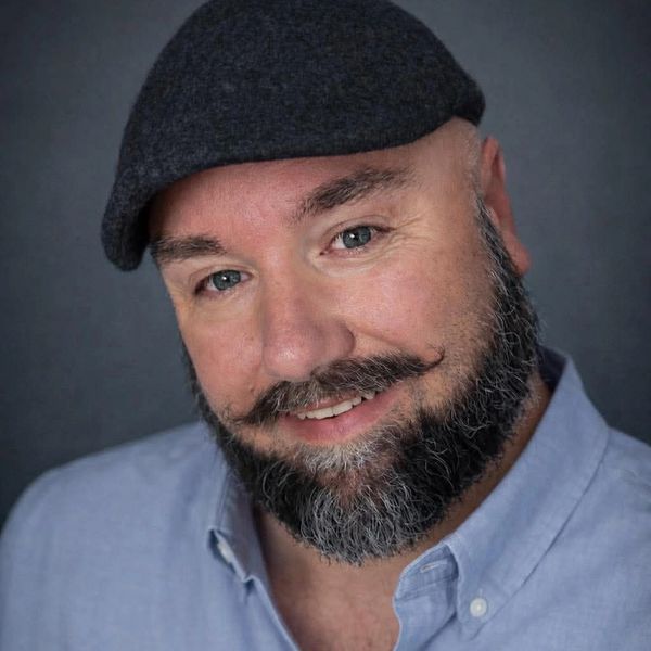 Smiling man with a beard and mustache wearing a cap and blue shirt.