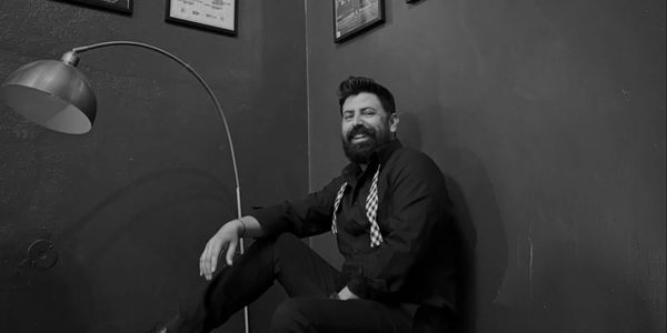 A bearded man in suspenders smiles while sitting by a lamp in a monochrome room.