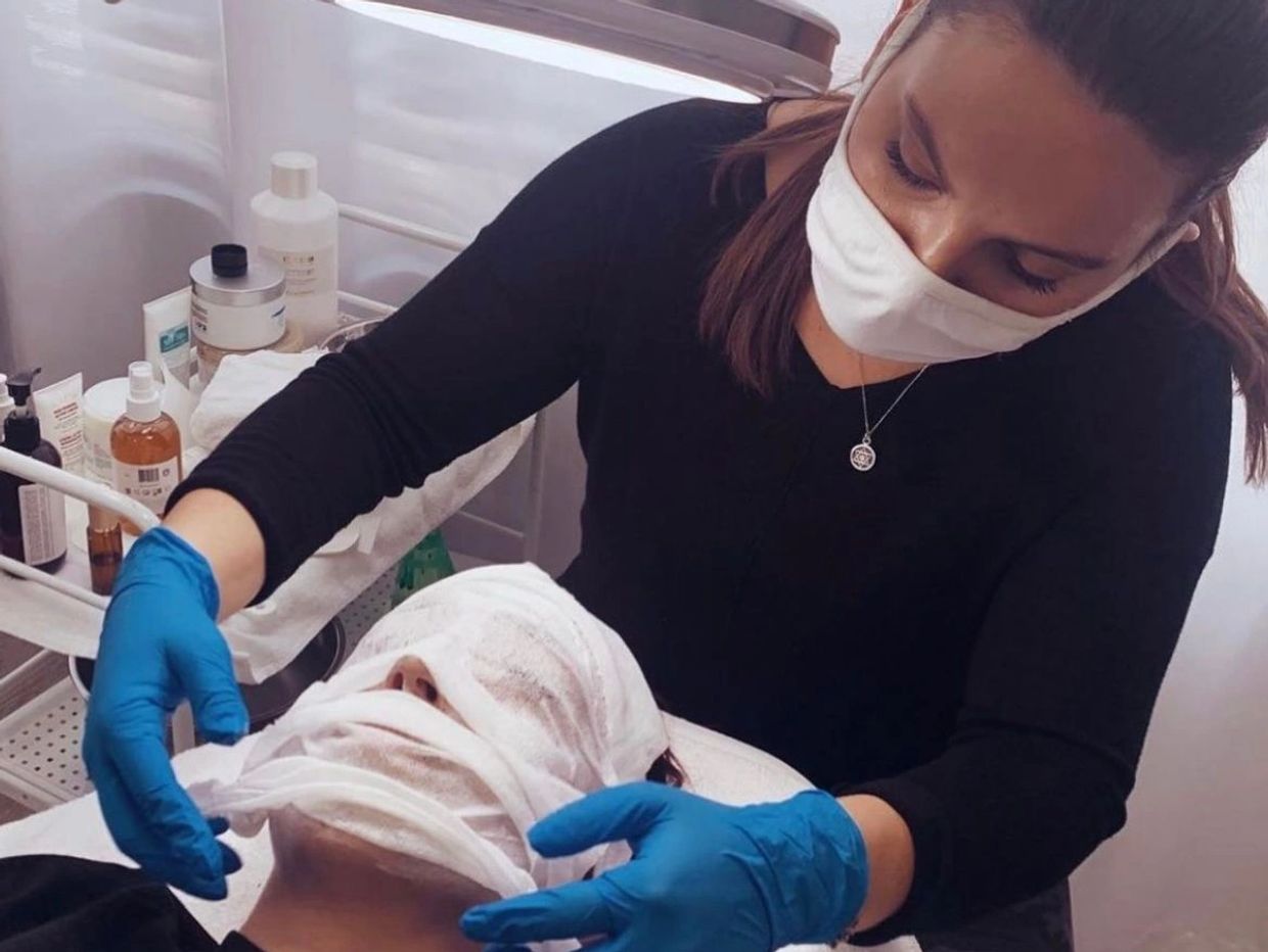 A beautician in blue gloves wraps a face mask on a client's face during a skincare treatment. From Skin So Fly