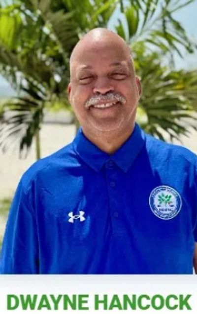 Smiling man in a blue polo shirt with a logo, standing outdoors with greenery behind him.