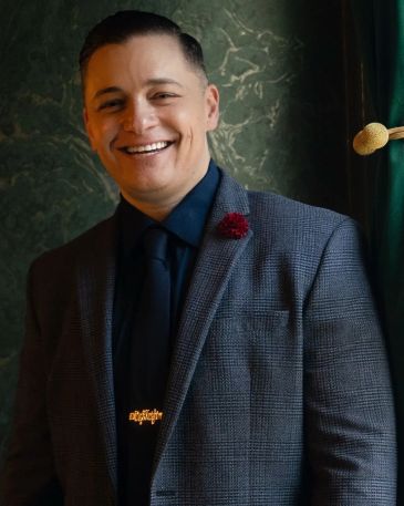 Smiling man in a checkered suit jacket and dark shirt with a red lapel pin.