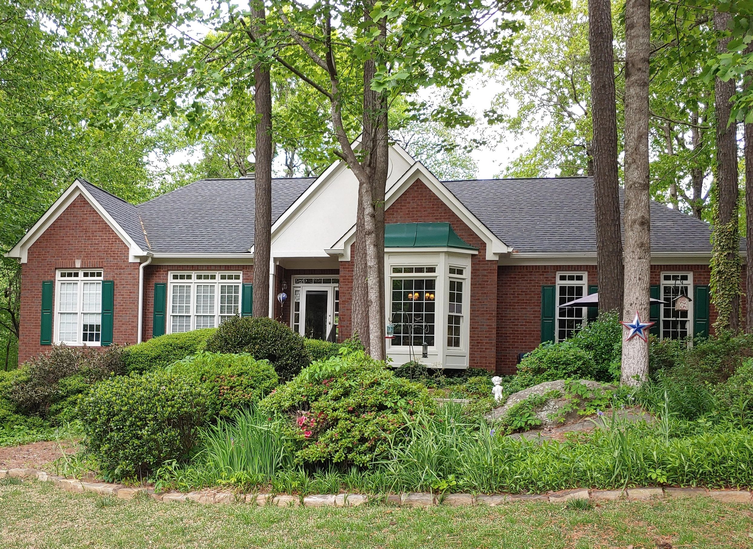 Brick house with green shutters surrounded by lush greenery and trees.