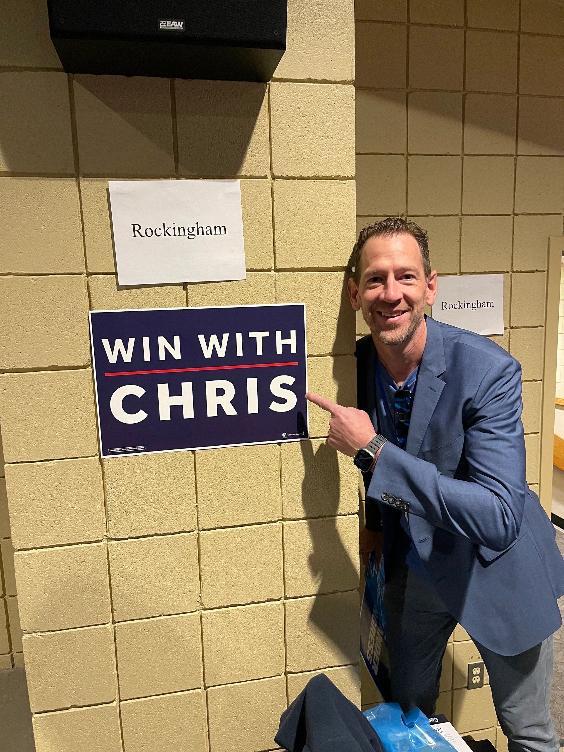 Man pointing at a 'Win with Chris' campaign sign indoors near a Rockingham sign.