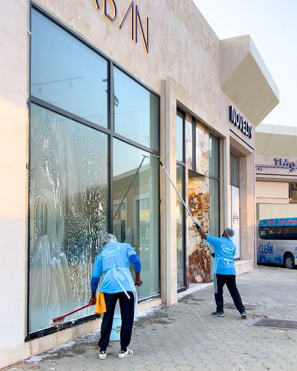 Two workers in blue uniforms cleaning large storefront windows on a sunny day offering cleaning services to our client