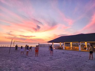 People playing beach volleyball at sunset with a colorful sky.