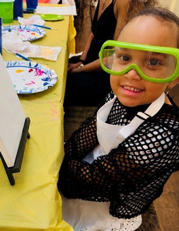Child wearing green safety goggles, apron, and black fishnet shirt at a painting table.