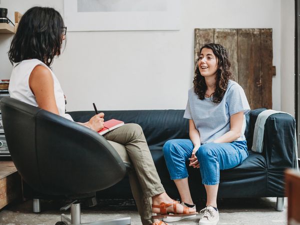 A therapist in a white top talks to a female patient in blue jeans on a dark sofa.