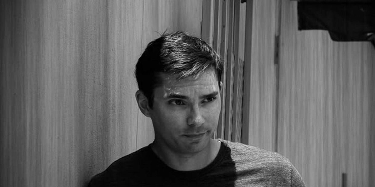 A man in a dark shirt leans against a wall in black and white.