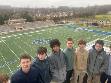 A group of teenage boys standing on bleachers overlooking a sports field on a cloudy day.