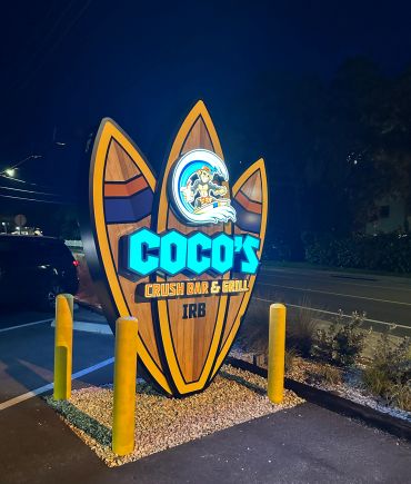 Illuminated surfboard-shaped sign for Coco's Crush Bar & Grill at night.