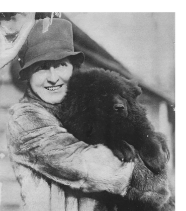 A middle-aged woman wearing a fur coat and a hat holds a black Chow dog in 1924.