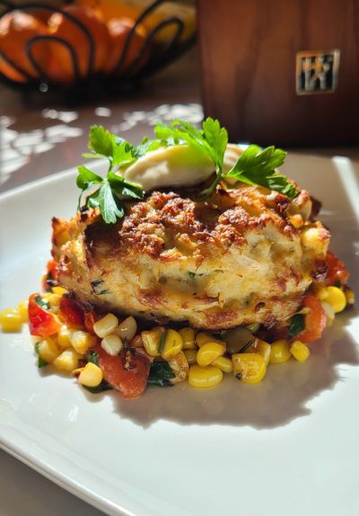 Golden crab cake served on a bed of sautéed corn and peppers, garnished with fresh parsley and a dollop of sauce.