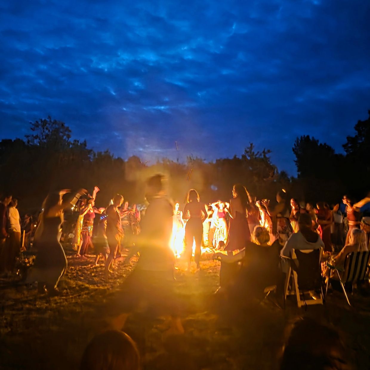 People dancing around a bonfire at night under a cloudy sky.
