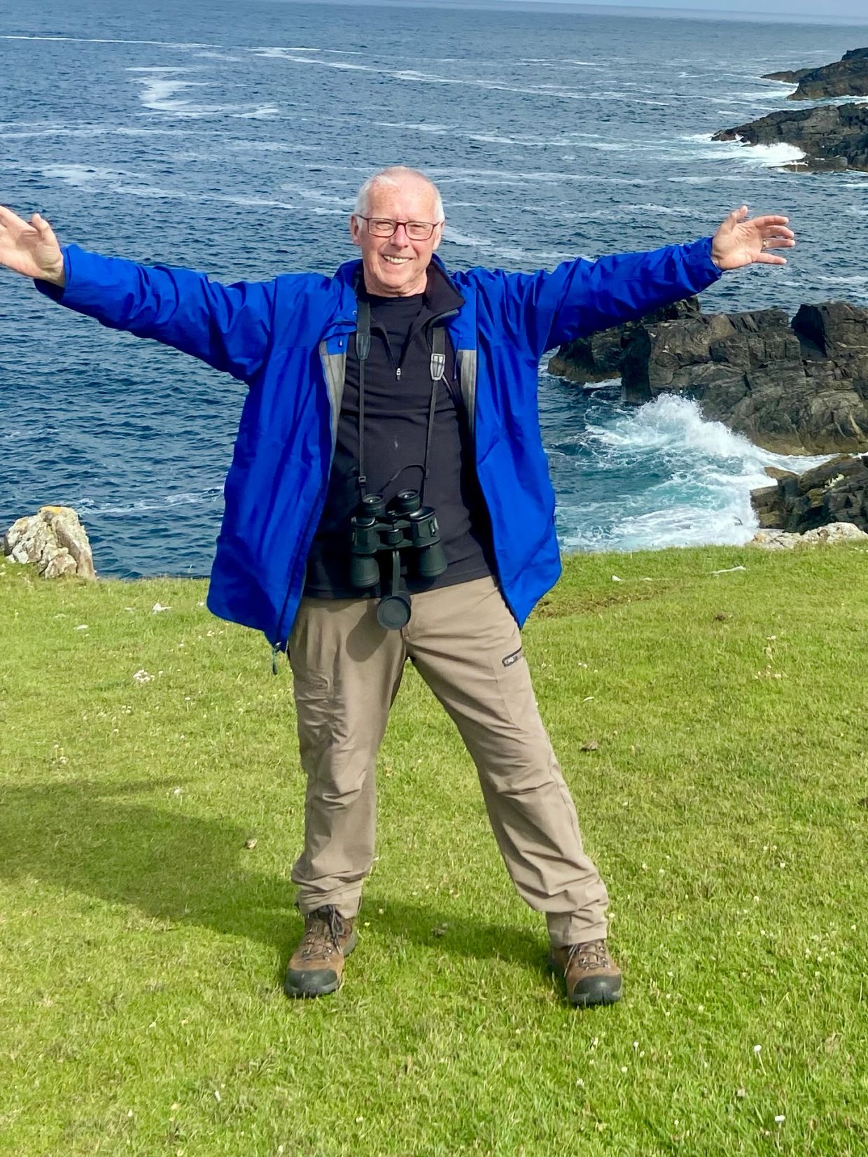 Man in blue jacket with binoculars near the ocean.