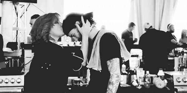 A woman kisses a man's forehead in a busy kitchen setting.