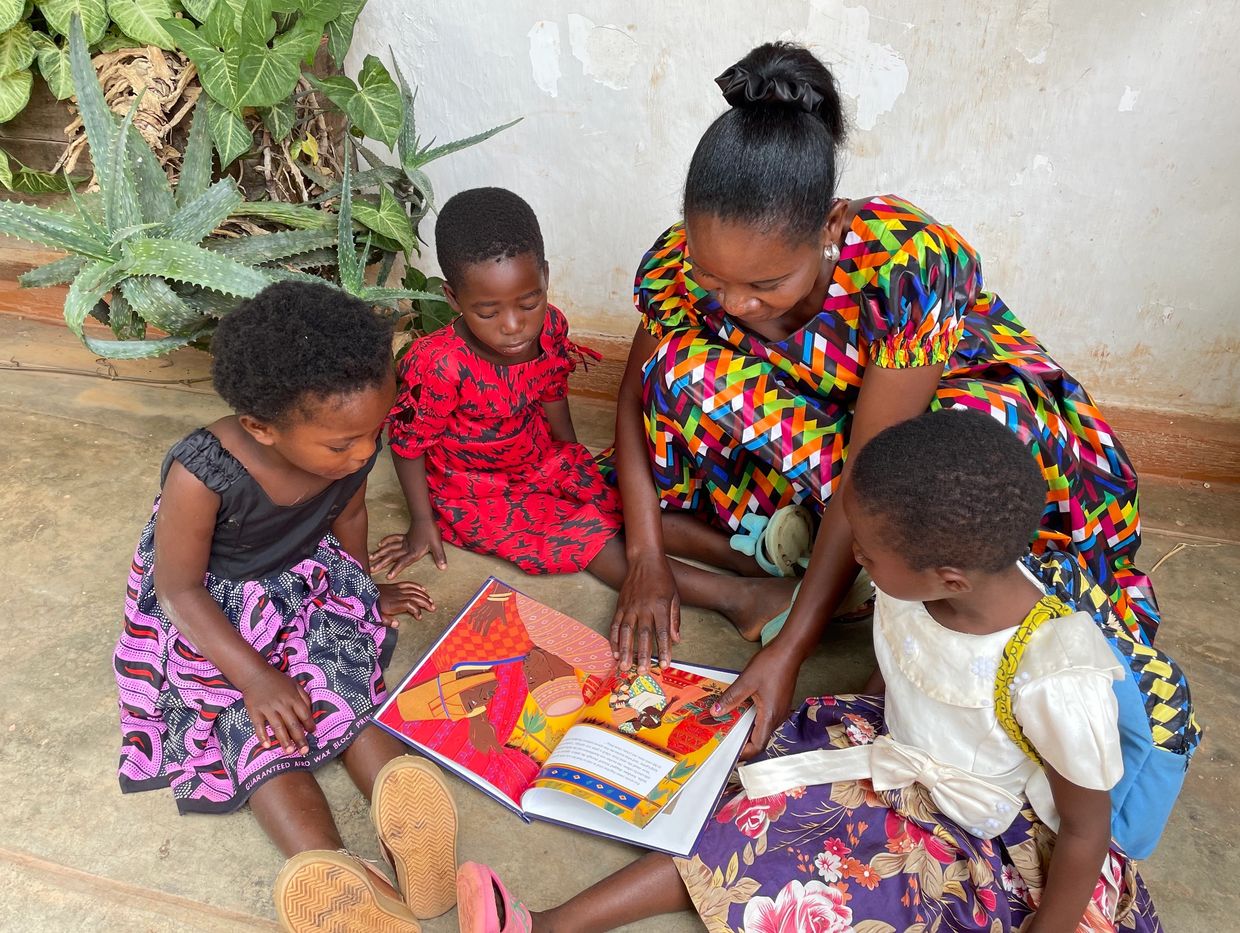 A woman reads a colorful book to three children sitting on the floor.