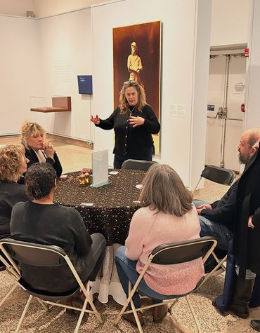 A woman leads a discussion with a seated group around a table in an art gallery.