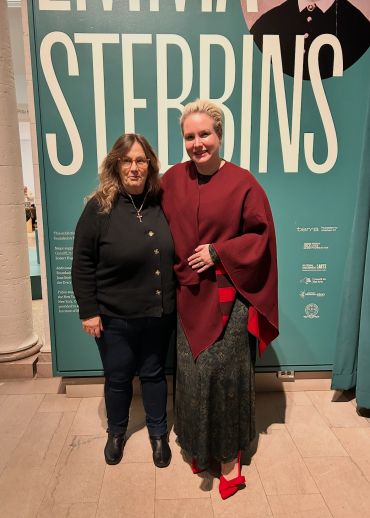 Two women posing together in front of a STEBBINS exhibition banner.