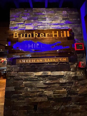 Rustic stone wall with a wooden sign for Bunker Hill American Taproom under blue lighting.